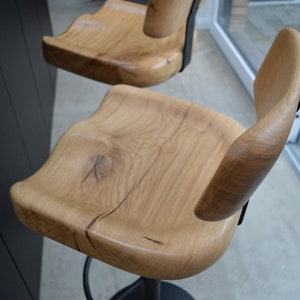 May include: A close-up of a wooden bar stool with a natural wood finish. The stool has a curved back and a wide, comfortable seat. The stool is made of solid wood and has a black metal base.