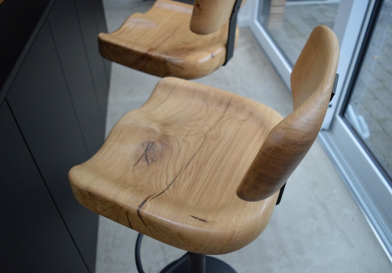 May include: A close-up of a wooden bar stool with a natural wood finish. The stool has a curved back and a wide, comfortable seat. The stool is made of solid wood and has a black metal base.