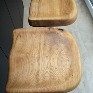 Sculpted oak counter height stool with a unique solid wood top and minimalist black metal base. Set of two designer counter stools accenting a sleek dark kitchen island with concrete flooring and rustic decor accents.