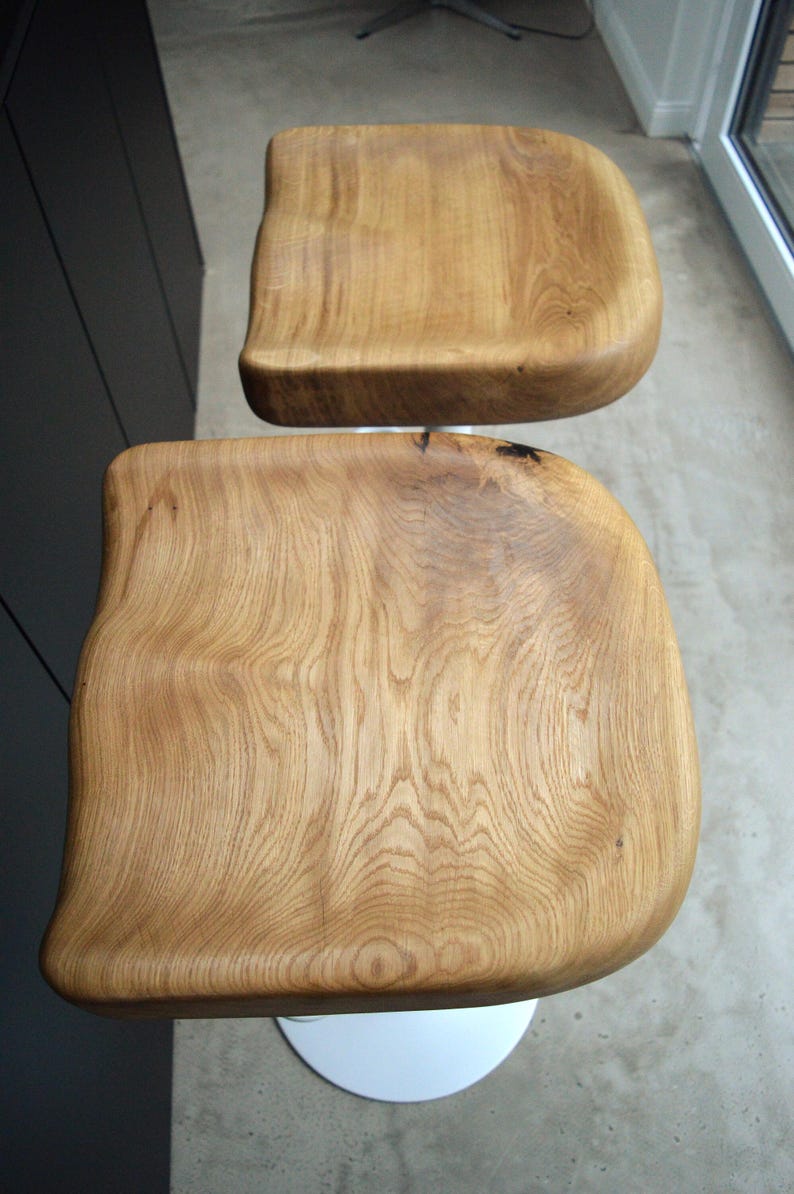 Sculpted oak counter height stool with a unique solid wood top and minimalist black metal base. Set of two designer counter stools accenting a sleek dark kitchen island with concrete flooring and rustic decor accents.