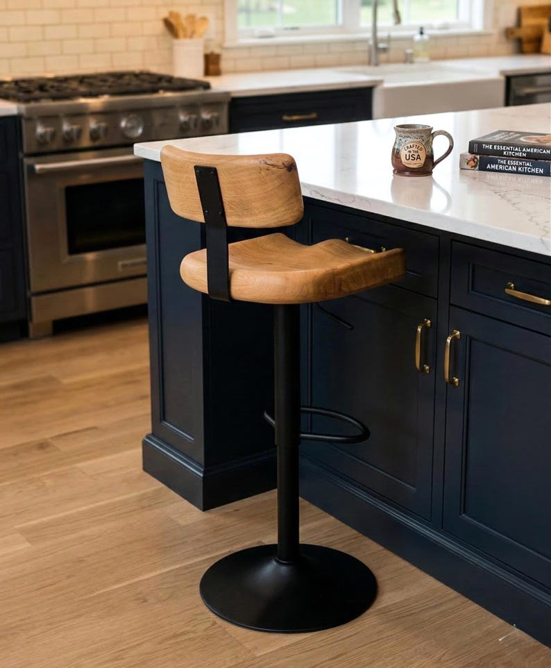 A contemporary, adjustable bar stool with a natural oak wood seat and backrest and a black pedestal base is positioned at a dark blue kitchen island with a white marble countertop. The background features a detailed high-end kitchen with a stainless steel range, subway tile backsplash, white cabinets, and a window looking out onto a garden. A mug and stacked cookbooks are visible on the island surface.