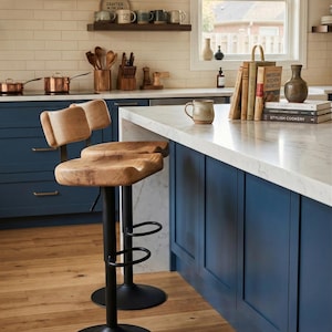Pair of handcrafted live edge oak bar stools with ergonomic tractor-style wooden seats and adjustable black metal pedestal bases, styled at a modern navy blue kitchen island with a white marble countertop in a bright, farmhouse-inspired American kitchen.