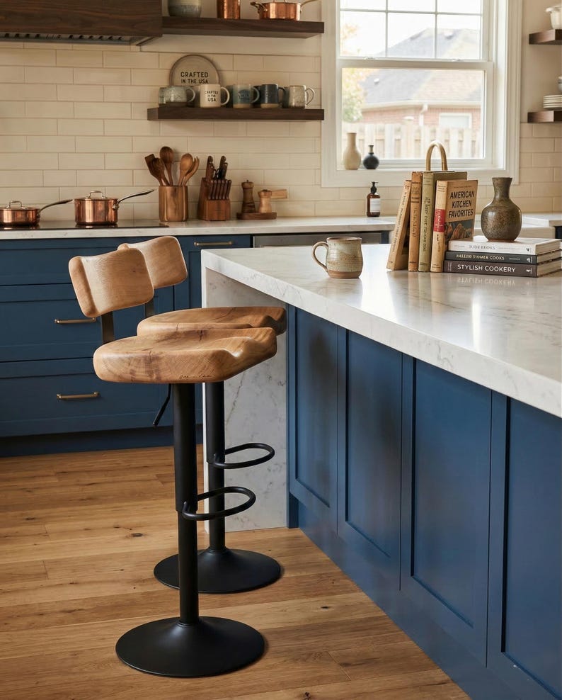 Pair of handcrafted live edge oak bar stools with ergonomic tractor-style wooden seats and adjustable black metal pedestal bases, styled at a modern navy blue kitchen island with a white marble countertop in a bright, farmhouse-inspired American kitchen.