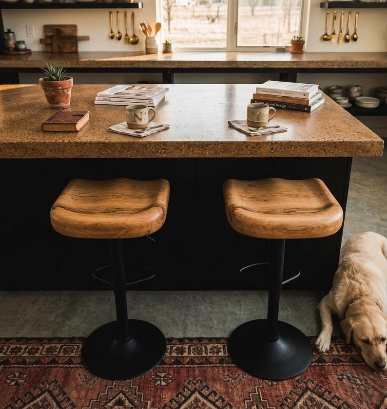 Premium wooden Bar stools, counter stool with an ergonomically carved solid oak seat showcasing luxury wood grain and natural character. Matte black metal swivel base positioned at a modern black kitchen island. High-end seating solution for organic modern and contemporary kitchen spaces.