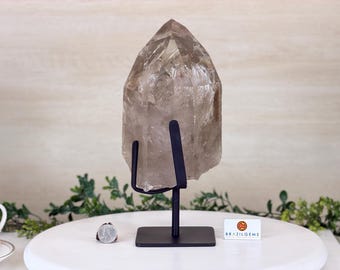 May include: A large, translucent smoky quartz crystal on a black metal stand. The crystal has a pointed top and a rough, natural texture. The stand has a square base and a curved support. The background is a neutral white with greenery.