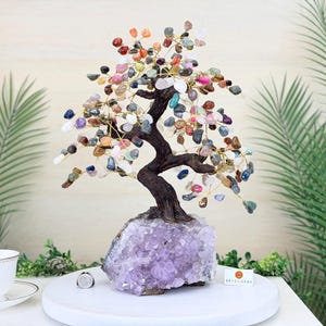 May include: A decorative tree sculpture featuring a dark brown textured trunk anchored to a large, jagged amethyst crystal base. The branches are made of thin gold-toned wire adorned with numerous small, polished multi-colored gemstone chips.