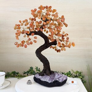 May include: A decorative tree sculpture with a dark brown, curved trunk and branches adorned with orange-colored gemstone leaves. The tree sits on a black base with purple crystals. The sculpture is a unique home decor item.