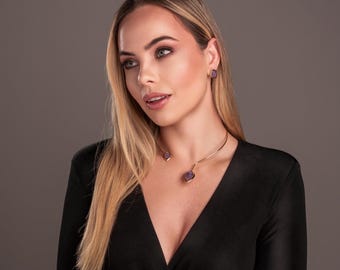 May include: A woman wearing a black dress with a gold choker necklace and matching earrings with purple stones.