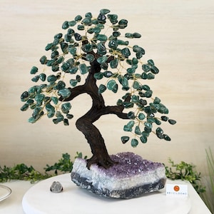 May include: A handcrafted bonsai tree sculpture with a dark brown trunk and branches, adorned with green gemstone leaves. The tree sits atop a purple and white amethyst base, creating a unique decorative piece.