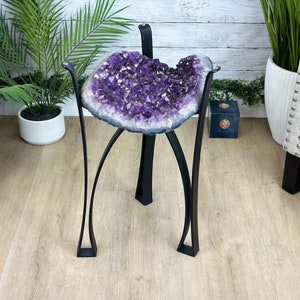 May include: A purple amethyst geode on a black metal stand. The geode is large and has a natural, rough texture. The stand has three legs and is made of sturdy metal.