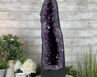 May include: A large amethyst geode with a deep purple color and a natural, rough exterior. The geode is displayed on a white pedestal.