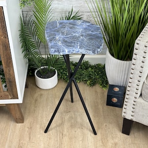 May include: A blue and white marble top side table with a black metal tripod base. The table is sitting on a wooden floor with a white cabinet and a gray sofa in the background.