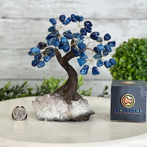 May include: A decorative tree sculpture with a brown trunk and branches, adorned with blue gemstone leaves. The tree sits on a white and gray crystal base. A silver ring and a box with the text "BRAZIL GEMS" are also present.