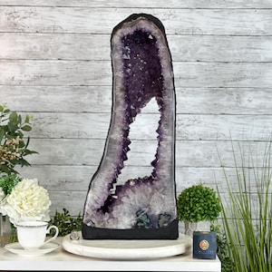 Può includere: Una grande geode di ametista con un'apertura naturale, che mostra i cristalli viola all'interno. La geode è esposta su un piedistallo bianco con una base nera.