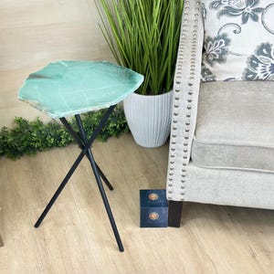 May include: A small, decorative side table with a light green, irregular-shaped top. The table has a black, tripod-style metal base. The table is near a couch and a potted plant. The table's top is approximately 18 inches in diameter.