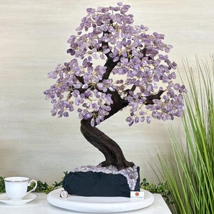 May include: A decorative amethyst tree sculpture with a dark brown trunk and branches, and purple amethyst leaves. The tree sits on a dark gray base and is displayed on a white surface. The sculpture is a unique home decor item.