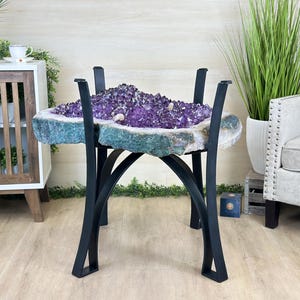 May include: A unique amethyst crystal table with a dark metal base. The amethyst features a vibrant purple center surrounded by a teal and white edge. The table's legs are black and curved, adding a modern touch to the natural stone.