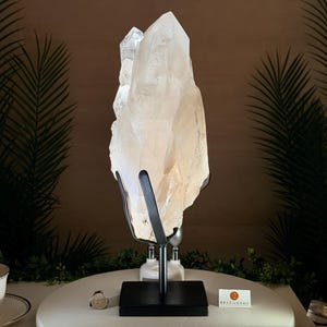 May include: A large, clear quartz crystal cluster displayed on a black metal stand. The crystal is illuminated, showcasing its natural formations. The stand is on a white surface, with a teacup and saucer visible in the foreground.