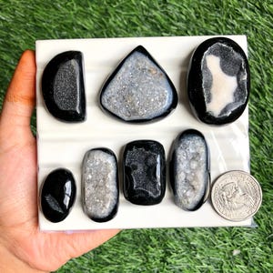May include: A collection of polished black and gray geode stones in various shapes, including oval, teardrop, and triangular. The geodes are displayed on a white surface, with a US quarter for size comparison. The stones have a sparkling, crystalline interior.