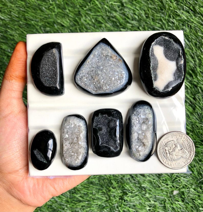 May include: A collection of polished black and gray geode stones in various shapes, including oval, teardrop, and triangular. The geodes are displayed on a white surface, with a US quarter for size comparison. The stones have a sparkling, crystalline interior.