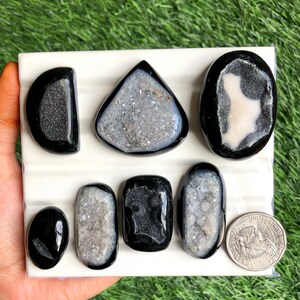 May include: A collection of polished black and gray geode stones in various shapes, including oval, teardrop, and triangular. The geodes are displayed on a white surface, with a US quarter coin for size comparison. The background is green.