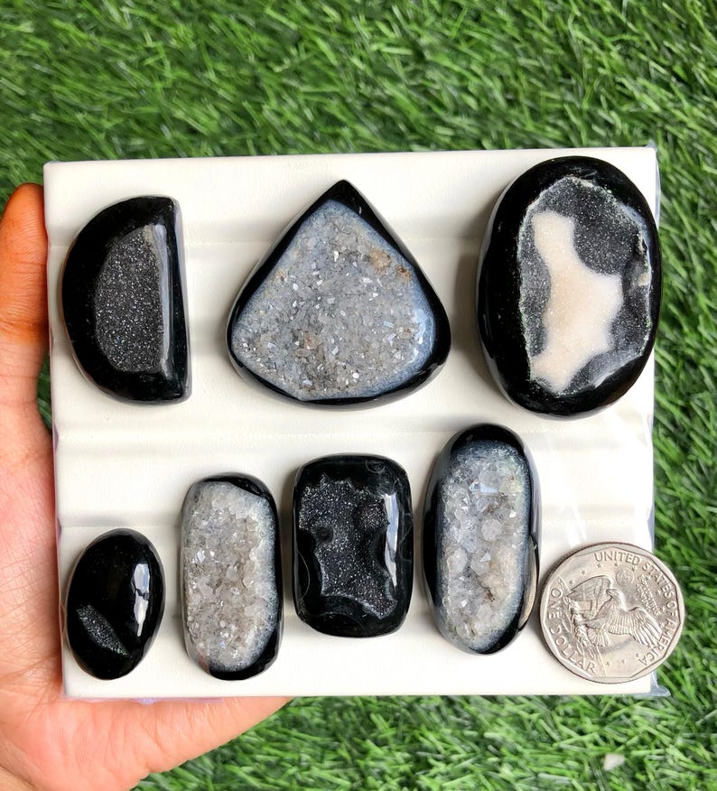 May include: A collection of polished black and gray geode stones in various shapes, including oval, teardrop, and triangular. The geodes are displayed on a white surface, with a US quarter coin for size comparison. The background is green.