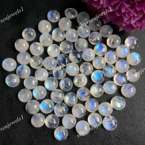 Könnte beinhalten: Eine Sammlung von weißen Mondstein-Cabochons mit schillernden blauen und grünen Blitzen. Die Steine sind oval geformt und haben unterschiedliche Größen. Die Steine sind auf einem schwarzen Hintergrund zu einem Haufen angeordnet.