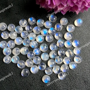 Könnte beinhalten: Eine Sammlung runder, polierter Mondstein-Cabochons. Die Steine sind durchscheinend mit einer milchig-weißen Basis und weisen eine blaue Adulareszenz auf. Die Edelsteine sind auf einer dunklen Oberfläche verstreut, mit ein paar rosa Blüten im Hintergrund.
