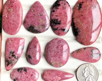 May include: A collection of polished rhodochrosite gemstones in various shapes, including ovals, teardrops, and rectangles. The stones display a vibrant pink hue with black and brown veining. A US quarter is included for size reference.