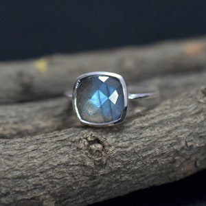 Puede incluir: Un anillo de plata con una piedra preciosa de labradorita de forma cuadrada. La piedra preciosa es de color azul y gris con destellos iridiscentes.