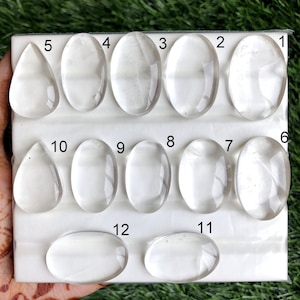 May include: A collection of twelve clear quartz gemstones in various shapes, including oval and teardrop. The gemstones are arranged on a white surface, with a hand holding the display against a green background.