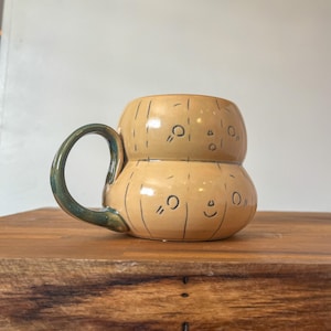 May include: A tan ceramic mug with a green handle. The mug has a stacked design with black line details and small, smiling faces. The mug is sitting on a wooden surface.