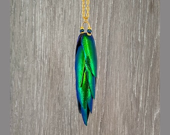 May include: A gold chain necklace with a pendant made from iridescent green beetle wings. The pendant is shaped like a teardrop and has a layered, textured look.