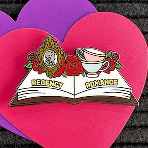 Op de afbeelding: Speld met een open boek met de woorden "Regency Romance", een theekopje, een ingelijst portret en rode rozen. De speld is geplaatst op een roze hartvorm over een paarse hartvorm. De achtergrond is een zwart, getextureerd oppervlak.