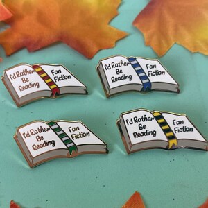 May include: Four enamel pins shaped like open books with the text "I'd Rather Be Reading" and "Fan Fiction". Each book has a different colored ribbon bookmark. The pins are on a light blue surface with fall leaves.