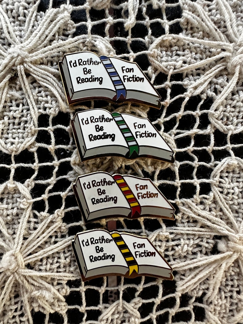 May include: Four enamel pins shaped like open books, each with the text "I'd Rather Be Reading" and "Fan Fiction." The book pins have different colored ribbon bookmarks: blue, green, red, and yellow. The pins are on a cream-colored, textured surface.
