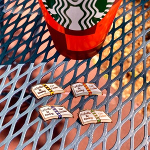 May include: Four enamel pins shaped like open books, each with the text "I'd Rather Be Reading Fan Fiction." The books have different colored ribbons and gold-colored borders. A Starbucks cup is in the background.