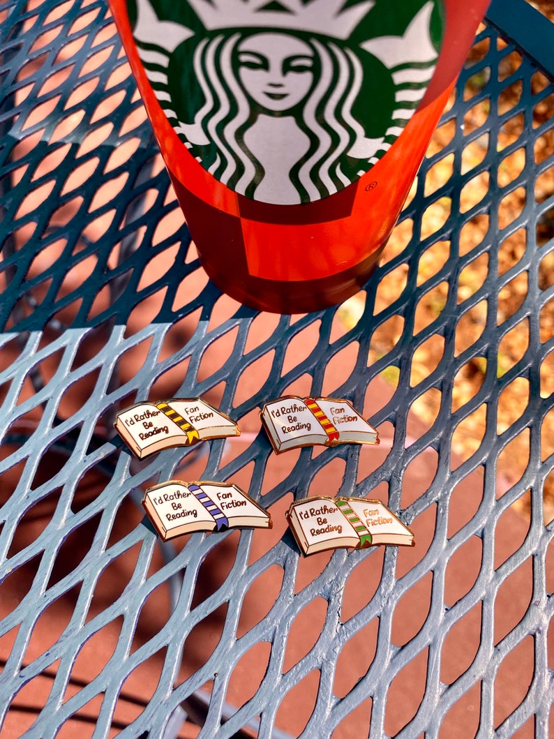 May include: Four enamel pins shaped like open books, each with the text "I'd Rather Be Reading Fan Fiction." The books have different colored ribbons and gold-colored borders. A Starbucks cup is in the background.