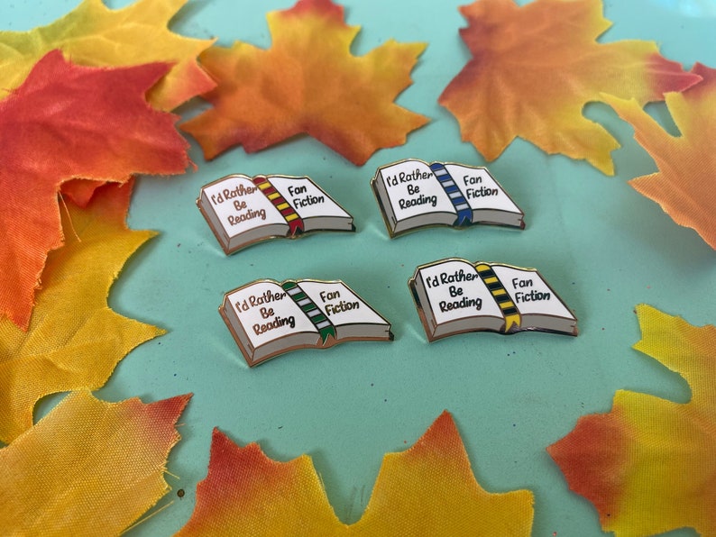 May include: Four enamel pins shaped like open books with the text "I'd Rather Be Reading" and "Fan Fiction". Each book has a different colored ribbon bookmark. The pins are on a light blue surface with fall leaves.