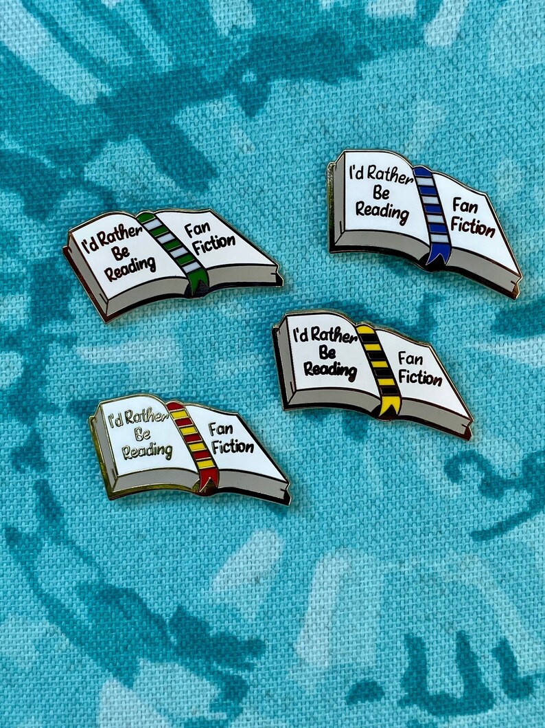 May include: Four enamel pins in the shape of open books, each with the text "I'd Rather Be Reading" and "Fan Fiction." The book spines are colored green, blue, yellow, and red. The pins have a gold border and are set against a teal patterned background.
