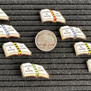 May include: Enamel pins in the shape of open books with the text "I'd Rather Be Reading" and "Fan Fiction". The pins have gold borders and colorful ribbon bookmarks. A Connecticut quarter is also in the image.