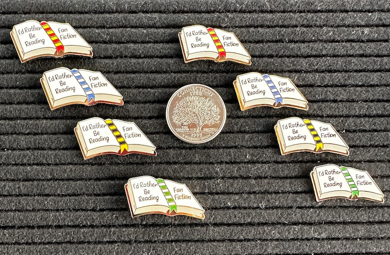 May include: Enamel pins in the shape of open books with the text "I'd Rather Be Reading" and "Fan Fiction". The pins have gold borders and colorful ribbon bookmarks. A Connecticut quarter is also in the image.