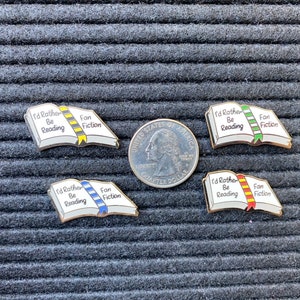 May include: Four enamel pins shaped like open books, each with the text "I'd Rather Be Reading" and "Fan Fiction." The pins have different colored ribbons: yellow, blue, green, and red. A quarter coin is next to the pins.