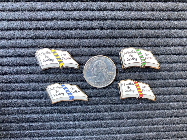 May include: Four enamel pins shaped like open books, each with the text "I'd Rather Be Reading" and "Fan Fiction." The pins have different colored ribbons: yellow, blue, green, and red. A quarter coin is next to the pins.