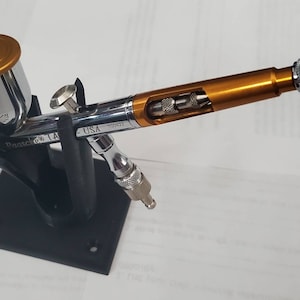 May include: An airbrush with a gold-colored body and a silver-colored nozzle. The airbrush is mounted on a black stand. The airbrush is labeled "Paasche" and "USA".
