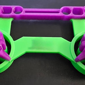 Peut inclure: Un support de manette imprimé en 3D en forme de crâne vert et violet. Le support comporte deux fentes en forme de crâne pour les manettes et une barre violette en haut.