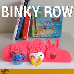 May include: Pink silicone pacifier holder with three animal faces: a cat, a bear, and a rabbit. Each animal has a slot for a pacifier. The holder is on a white surface.