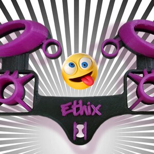 May include: A 3D printed black and purple phone holder with the word "Ethix" printed on the front. The holder has two rings on the top and two circular openings on the bottom. A yellow emoji with a tongue sticking out is in the background.