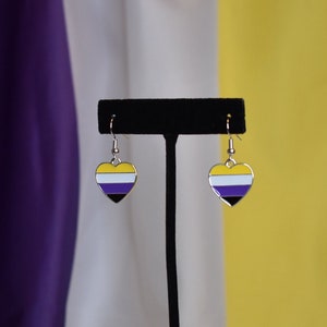 May include: A pair of silver heart-shaped earrings with a yellow, white, purple, and black striped design. The earrings are displayed on a black stand.
