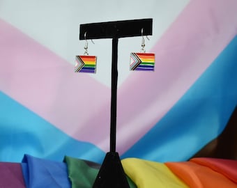 May include: A pair of silver dangle earrings featuring a rainbow flag design. The earrings are displayed on a black stand against a backdrop of pink, blue and yellow fabric.